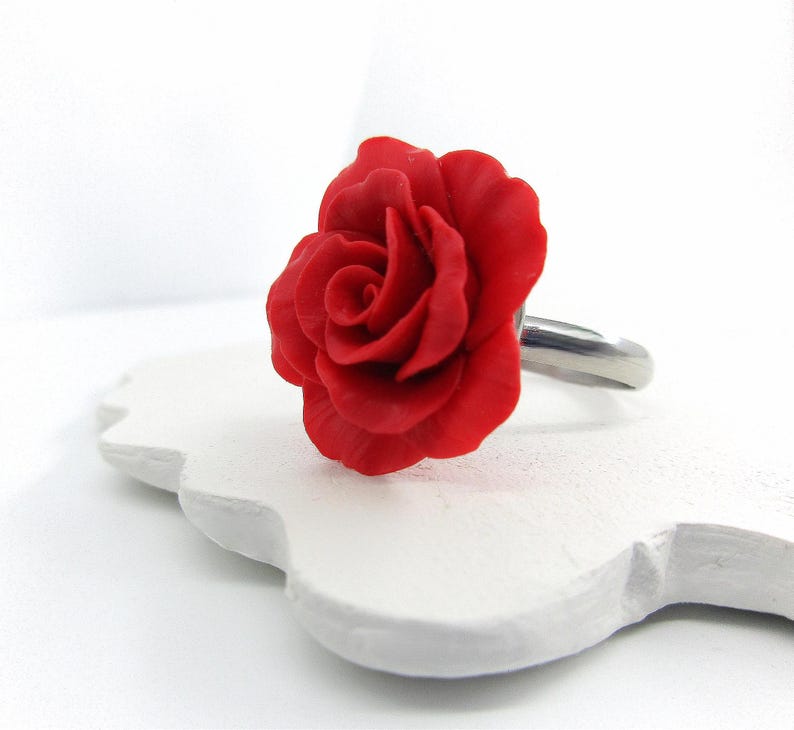 May include: A red rose ring with a detailed floral design. The rose is the focal point, set on a silver-colored band. The ring is displayed on a white, decorative surface.