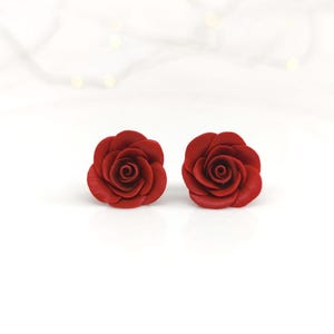 Op de afbeelding: Een paar oorknopjes in de vorm van dieprode rozen. Elke oorbel heeft een sculpturaal bloemendesign met gelaagde bloemblaadjes die in een spiraalpatroon zijn gerangschikt, wat een realistisch bloei-effect creëert. De oorbellen hebben een matte afwerking.