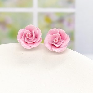 Könnte beinhalten: Ein Paar Ohrstecker in Form von hellrosa Rosenblüten. Jeder Ohrstecker ist mit geschichteten Blütenblättern geformt, die sich zur Mitte hin spiralförmig drehen, was ein realistisches florales Aussehen erzeugt. Die Ohrstecker sind als Set.
