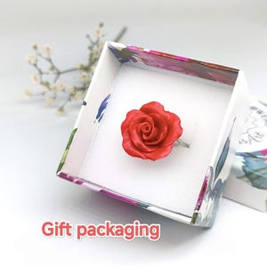 May include: A red rose ring displayed in a floral patterned gift box. The ring features a detailed, three-dimensional rose design. The box is white inside and has a floral pattern on the outside. The words "Gift packaging" are visible.