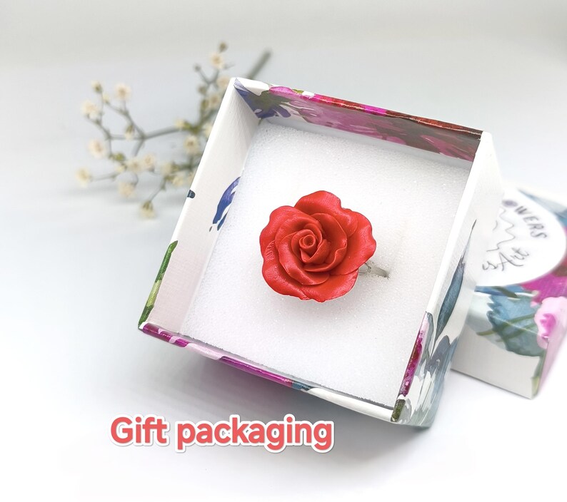 May include: A red rose ring displayed in a floral patterned gift box. The ring features a detailed, three-dimensional rose design. The box is white inside and has a floral pattern on the outside. The words "Gift packaging" are visible.