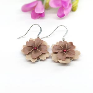 May include: A pair of flower-shaped earrings. Each earring features a layered design with petals in shades of beige and brown, centered with a small red bead. The earrings hang from silver-colored hooks, set against a white background.
