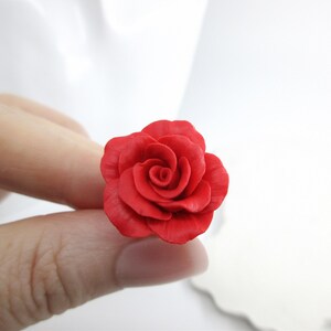 May include: A vibrant red rose, meticulously crafted, is held between fingers against a soft white background. The rose's petals are detailed, showcasing a realistic floral design. The image highlights the rose's rich color and texture.