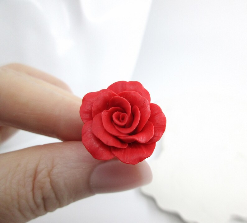 May include: A vibrant red rose, meticulously crafted, is held between fingers against a soft white background. The rose's petals are detailed, showcasing a realistic floral design. The image highlights the rose's rich color and texture.