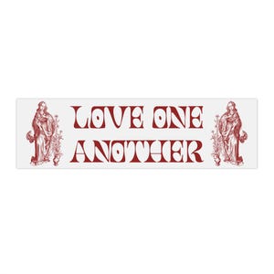 May include: A rectangular white sticker with the words "LOVE ONE ANOTHER" in red, stylised lettering. The sticker features a red illustration of a robed figure on each end, with floral accents.