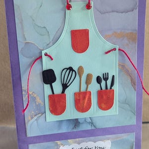 Puede incluir: Tarjeta de felicitación hecha a mano con un diseño de delantal azul claro, bolsillos rojos y utensilios de cocina. La tarjeta incluye el texto "Just for You" en una etiqueta blanca, sobre un fondo veteado y un marco morado.