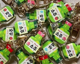 May include: A collection of clear plastic charms shaped like bottles with Korean text and images of fruit. The charms are attached to key rings and are likely used as decorative keychains.