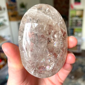 May include: A clear quartz crystal egg with iridescent inclusions. The egg is smooth and polished, and it is held in a hand.