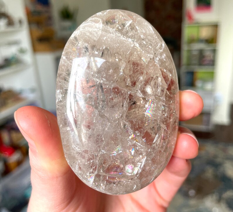 May include: A clear quartz crystal egg with iridescent inclusions. The egg is smooth and polished, and it is held in a hand.