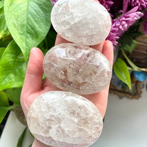May include: Three clear quartz crystal palm stones. The stones are smooth and have a cloudy, milky appearance.