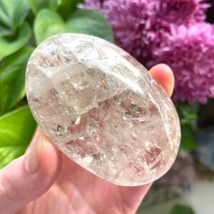 May include: A clear quartz crystal palm stone with a unique, textured surface. The crystal is smooth and polished, with a slightly oval shape.
