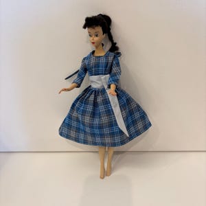以下が含まれることがあります： ポニーテールに黒髪のヴィンテージドール。人形は、白いサッシュが付いた青、グレー、白のチェック柄のドレスを着ています。ドレスは七分袖で、フレアスカートです。人形は立っています。