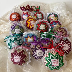 May include: A selection of handmade Christmas baubles, each with a star design and the year embroidered on the front. The baubles are in various colours, with white lace trim and colourful ribbons.
