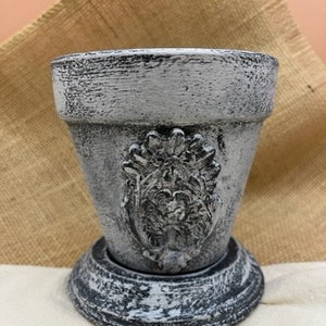 May include: A gray, distressed-finish flower pot with a decorative face design. The pot has a wide rim and sits on a matching base. The pot is made of a textured material, and the design is in a classical style.