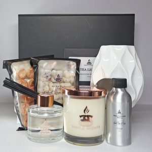 Op de afbeelding: Een cadeauset voor geur in huis met een witte keramische diffuser, een kaars met een koperen deksel, een flesje rietdiffuserolie en twee zakjes sojawas smelttabletten. Alle producten zijn gelabeld met de merknaam "Simple Scents" en de geur "Velvet Rose".