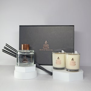Op de afbeelding: Een zwarte cadeaudoos met de tekst "Simple Scents" op de voorkant. De doos bevat twee glazen kaarsenpotten met witte was en een koperkleurig logo. Er is ook een glazen diffuserfles met een koperkleurig deksel en zwarte diffuserstokjes.