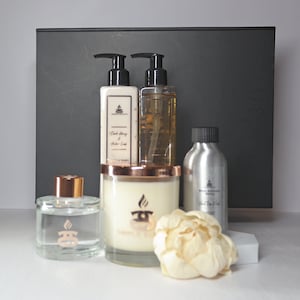 May include: A set of home fragrance products in a black box. The set includes a candle, a diffuser, a bottle of hand lotion, and a bottle of body wash. The products are all in clear glass bottles with gold accents. The candle is in a clear glass jar with a gold lid. The diffuser is in a clear glass bottle with a gold cap. The hand lotion and body wash are in clear glass bottles with gold pumps. The products are all labeled with the same brand name.