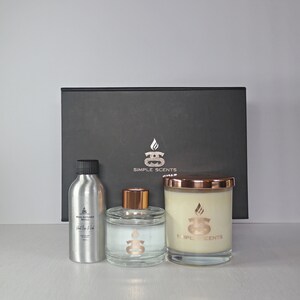 Puede incluir: Una caja de regalo negra con un logotipo dorado que dice "Simple Scents" contiene una vela, un difusor y una botella de aceite esencial. La vela y el difusor tienen tapas doradas.