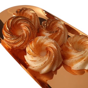 May include: Four swirl-shaped, light brown, cake-like treats with gold flecks on a copper-coloured tray.
