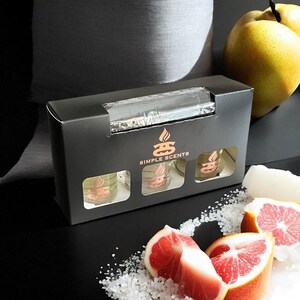 May include: A black box with the text "SIMPLE SCENTS" on the front. The box contains three small glass jars with a gold lid. The jars are filled with a clear liquid. The box is sitting on a black surface with a grapefruit, salt, and a pear.