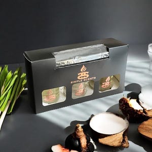 May include: A black box with three clear glass jars containing scented wax melts. The box has the text "Simple Scents" printed on the front. The jars are arranged in a row and are partially visible through the box's opening.