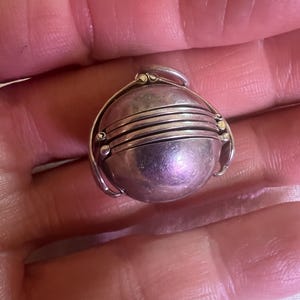 Puede incluir: Un medallón esférico plateado con un diseño de bisagra. El medallón presenta múltiples bandas horizontales y pequeños detalles decorativos. El colgante redondo se sostiene en una palma abierta, mostrando su tamaño y detalles.