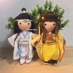 Può includere: Due bambole all'uncinetto, una con un vestito bianco e blu con una fascia blu e l'altra con un vestito giallo con un cappello marrone. Le bambole sono in piedi su una superficie di legno.