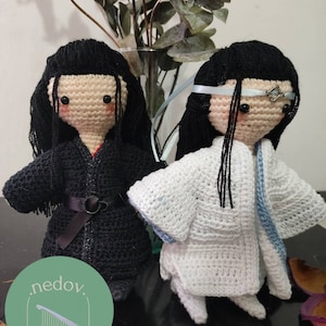 May include: Two crocheted dolls wearing traditional Chinese clothing. The doll on the left is wearing a black robe with a black belt. The doll on the right is wearing a white robe with a blue sash. Both dolls have black hair and brown eyes.