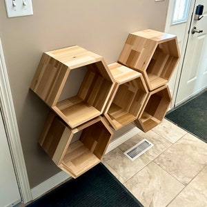 May include: A set of six wooden hexagonal shelves, arranged in a honeycomb pattern, mounted on a wall. The shelves are made of light brown wood and have a natural finish.