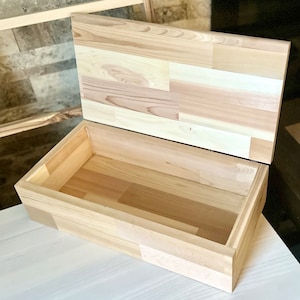 May include: A wooden box with a hinged lid, made of light-coloured wood with a natural grain pattern. The lid is open, revealing the empty interior of the box.