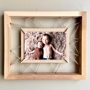 May include: A wooden picture frame with a photo of two children. The frame is made of light-colored wood and has a unique design with twine strung across the opening. The photo features a baby wrapped in a brown blanket and an older child.