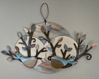 May include: A metal wall art piece featuring two stylized birds perched on branches with leaves. The birds are painted in shades of blue and brown, and the branches are painted in brown. The background is a circular design with a white and brown paint effect.