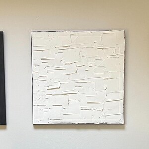 May include: Three square canvases with black and white abstract designs. The centre canvas is white with a textured surface. The canvases on either side are black with a pattern of small raised dots.