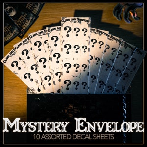 Może przedstawiać: Czarna koperta z tekstem "MYSTERY ENVELOPE 10 ASSORTED DECAL SHEETS" na przodzie. Koperta jest otwarta i zawiera dziesięć arkuszy białego papieru z czarnymi znakami zapytania wydrukowanymi na nich. Każdy arkusz ma wydrukowany tekst "CLAWS AND STILETTOS NAIL DECALS".