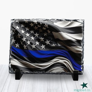 May include: A slate plaque with a black and white American flag with a blue line running horizontally across the flag. The flag is waving in the wind. The plaque is on a wooden surface.