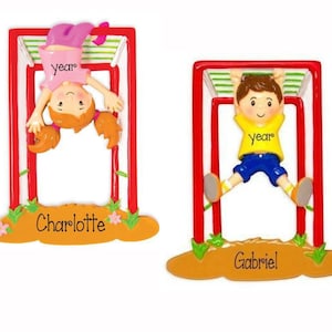 May include: Two personalized Christmas ornaments featuring children hanging from a red and white jungle gym. The girl's ornament is red, white, and pink with the name Charlotte. The boy's ornament is red, white, and blue with the name Gabriel.