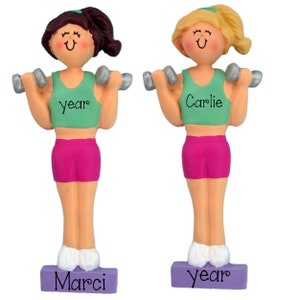 May include: Two personalized ornaments of women lifting weights. The women are wearing green tank tops with the names "Marci" and "Carlie" on them. The ornaments are on purple bases with the word "year" on them.