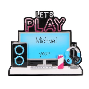 May include: A gaming-themed ornament featuring a computer monitor with the text Michael and year, a black speaker with blue accents, a keyboard, a pink and white beverage can, and blue headphones. The top displays the text LET'S PLAY in pink.