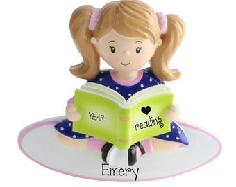 May include: A figurine of a child with light brown hair in pigtails, wearing a blue dress with white polka dots, sitting on an oval base. The child holds a lime green open book with the text YEAR reading and a black heart. The name Emery is printed on the base.