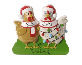 May include: A decorative ornament featuring two chickens standing on a green base. The brown chicken wears a red and white scarf labeled Rudy, and the white chicken wears a string of colorful lights labeled Girtie. Both wear Santa hats with text year Farm Living.
