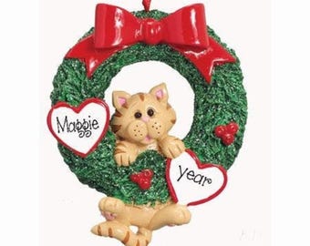 May include: A green glittery wreath ornament featuring a tan tabby cat in the center. A large red bow sits at the top. Two white heart-shaped plaques are attached to the wreath, with the name Maggie on the left and the word year on the right.
