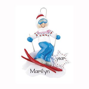 May include: A Christmas tree ornament featuring a skier wearing a white sweater with red hearts, blue pants, and a red and white hat. The skier holds two poles and stands on red skis. The base is white with the name Marilyn and a snowflake with the word year.