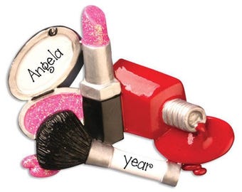 May include: A decorative ornament featuring a pink glittery lipstick, a black makeup brush with the word year printed on the handle, and a red nail polish bottle with spilled polish. An open compact case displays the name Angela in black script.