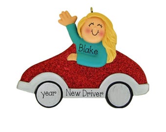 May include: A decorative ornament featuring a blonde character waving from a red glittery car. The character wears a teal shirt with the name Blake printed on it. The car has white fenders and wheels, with the text year New Driver printed on the side.
