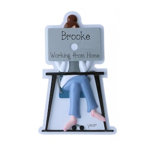 May include: A decorative ornament featuring a figure with brown hair in a bun sitting at a black desk. The figure holds a gray laptop with the text "Brooke" and "Working from Home" printed on it. The figure wears blue pants and the word "year" is printed below.