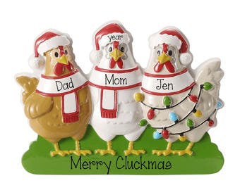 May include: A decorative ornament featuring three chickens standing on a green base with the text Merry Cluckmas. Each chicken wears a red and white Santa hat and a scarf labeled Dad, Mom, and Jen. The third chicken is wrapped in colorful lights.