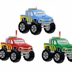 May include: Three monster truck ornaments in blue, red, and green. Each truck features large black tires, yellow side stripes, silver bumpers, and a roof light bar. The windows are personalized with the names Jaxon, Grayson, and Hudson, plus the word year.