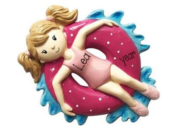 May include: A decorative ornament featuring a light-skinned figure with blonde hair in pigtails, wearing a pink swimsuit, floating inside a pink polka-dot inner tube surrounded by blue water splashes. The text Lea is on the swimsuit and year is on the tube.