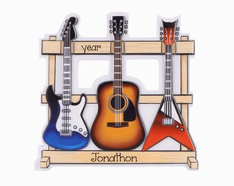 May include: A decorative ornament featuring three guitars on a wooden rack. The left is a blue and white electric guitar, the center is a brown acoustic guitar, and the right is a red and black V-shaped electric guitar. The text year and Jonathon appear.
