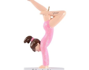 May include: A gymnast ornament wearing a glittery pink leotard, performing a handstand with one leg extended upward. The figure has brown hair in a ponytail, features the word year on the leg, and stands on a white base labeled with the name Christina.